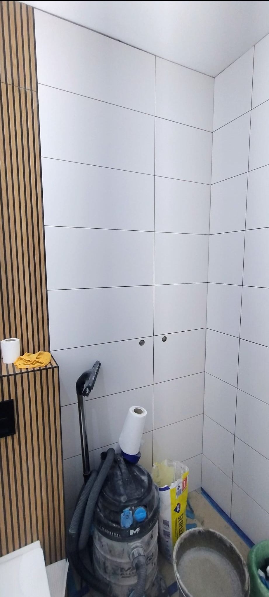 Białe, prostokątne płytki ścienne w łazience, częściowo wykończone, z widocznymi otworami na armaturę. Obok ściany z drewnianymi lamelami stoi odkurzacz i akcesoria.