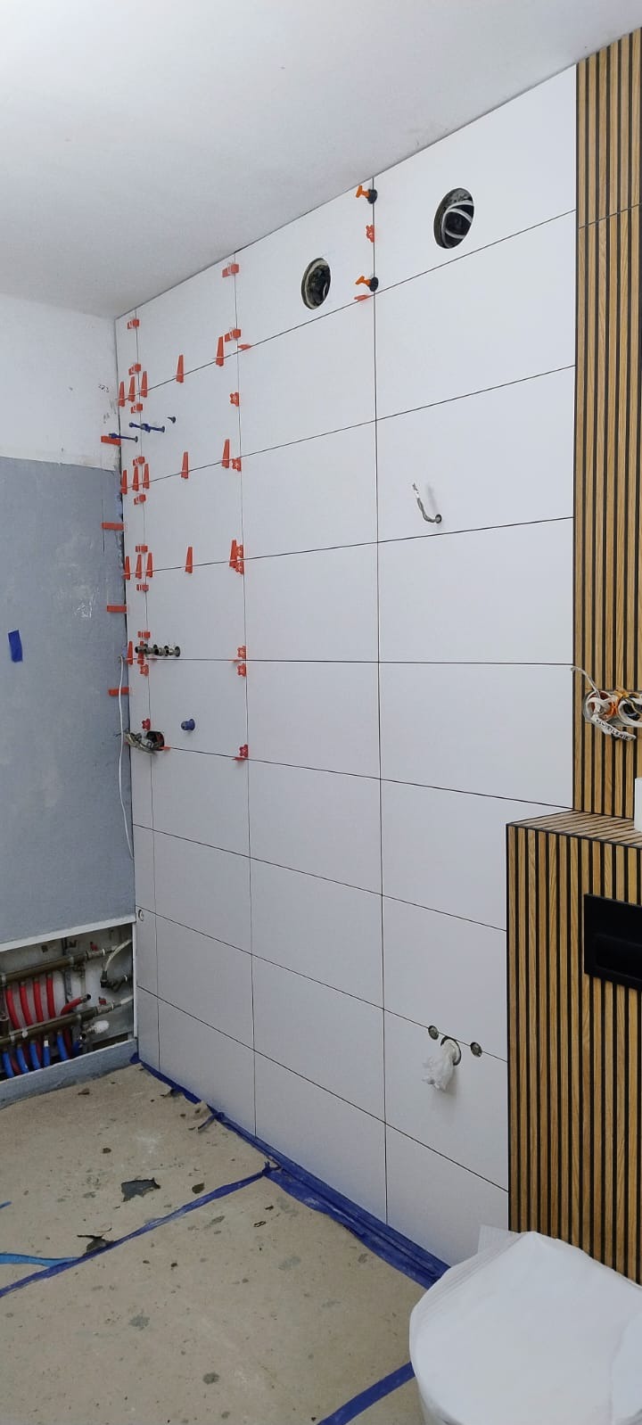 Białe płytki ścienne w trakcie montażu w łazience, z widocznymi systemami poziomowania i fragmentem obudowy z drewnianych listew. Widoczne instalacje wodne.