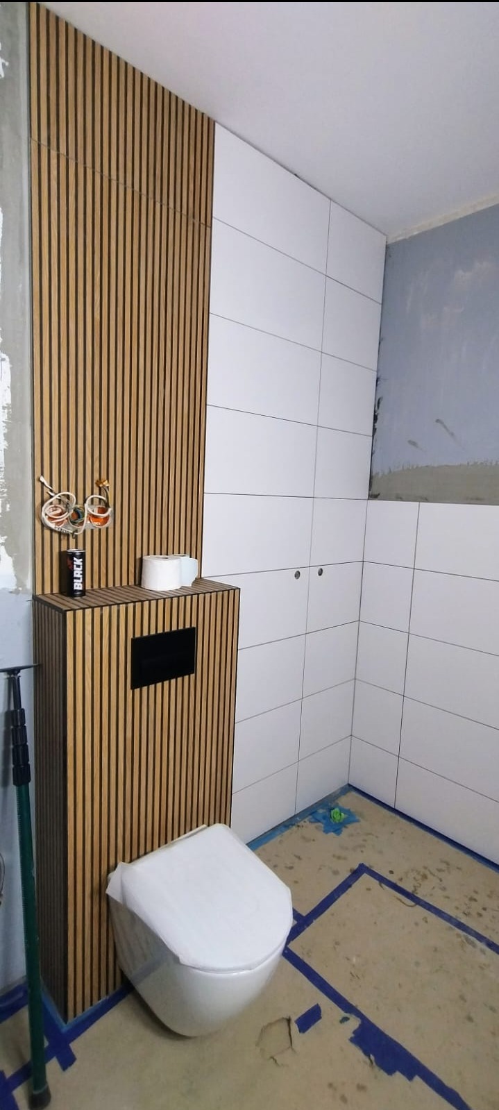 Nowoczesna łazienka w trakcie wykończenia: białe płytki, drewniane lamele, podwieszana toaleta. Widoczne elementy instalacji i zabezpieczona podłoga.