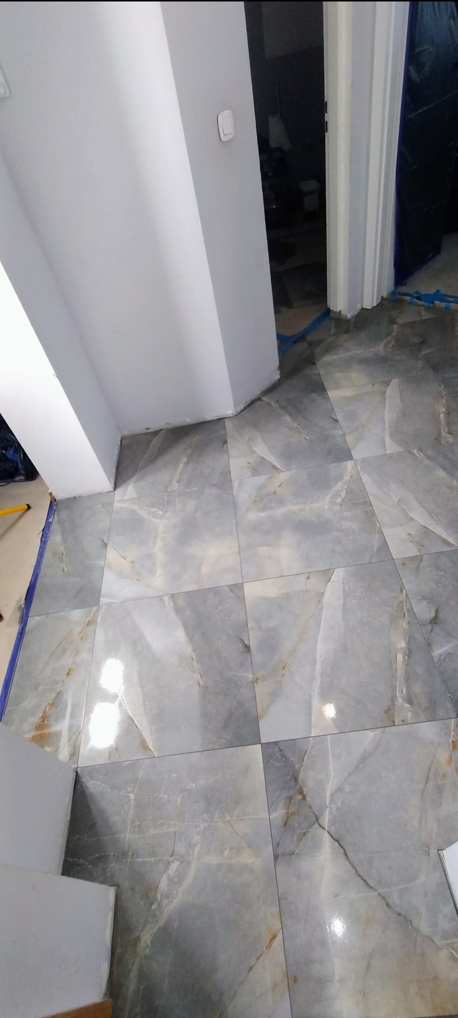 Ułożone, błyszczące płytki podłogowe imitujące marmur w odcieniach szarości i beżu, widok z góry, fragment ściany i futryny drzwiowej.