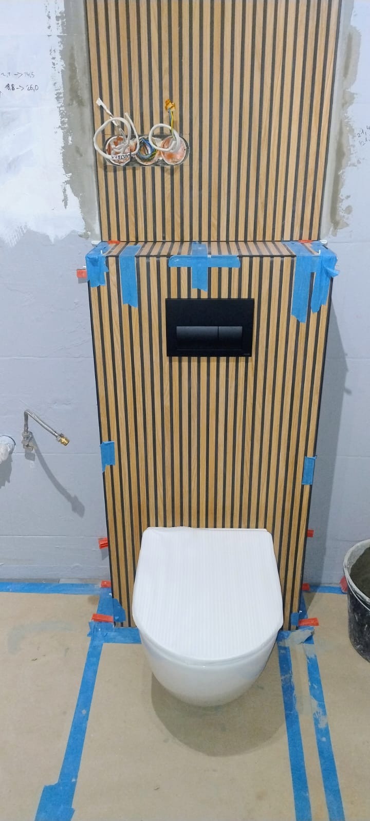 Podwieszana toaleta z obudową z drewnianych lameli w trakcie montażu. Widoczne taśmy maskujące, poziomowanie i wystające przewody elektryczne.