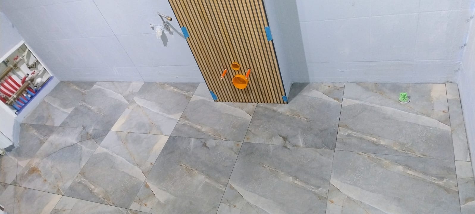 Wykończona łazienka z szarymi płytkami imitującymi marmur na podłodze i ścianach. Widoczne drewniane lamele na ścianie i instalacja wodno-kanalizacyjna.