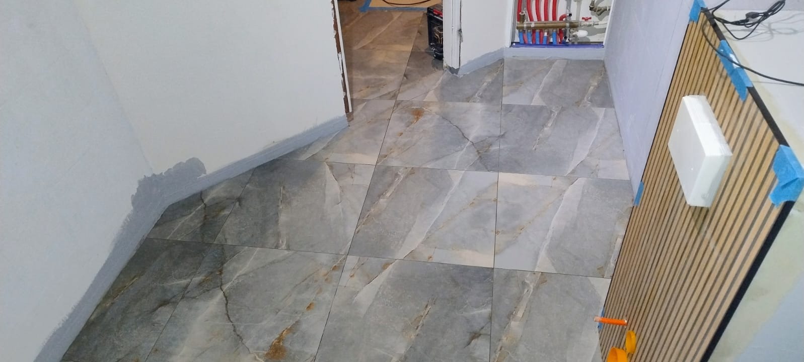 Ułożone szare płytki podłogowe z marmurkowym wzorem w korytarzu. Widoczne fragmenty ścian z szarą listwą przypodłogową i instalacja grzewcza. Częściowo widoczna mata grzewcza.