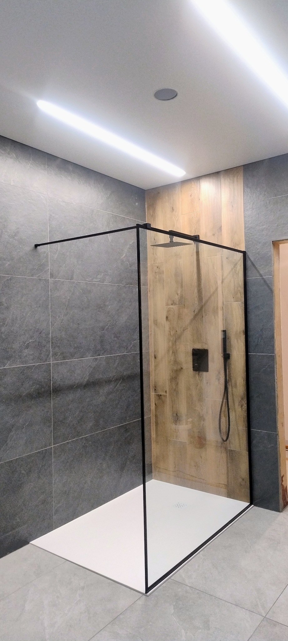 Nowoczesna kabina prysznicowa walk-in z czarnymi profilami, biały brodzik, szare płytki na ścianach i drewnopodobne panele. Oświetlenie sufitowe.