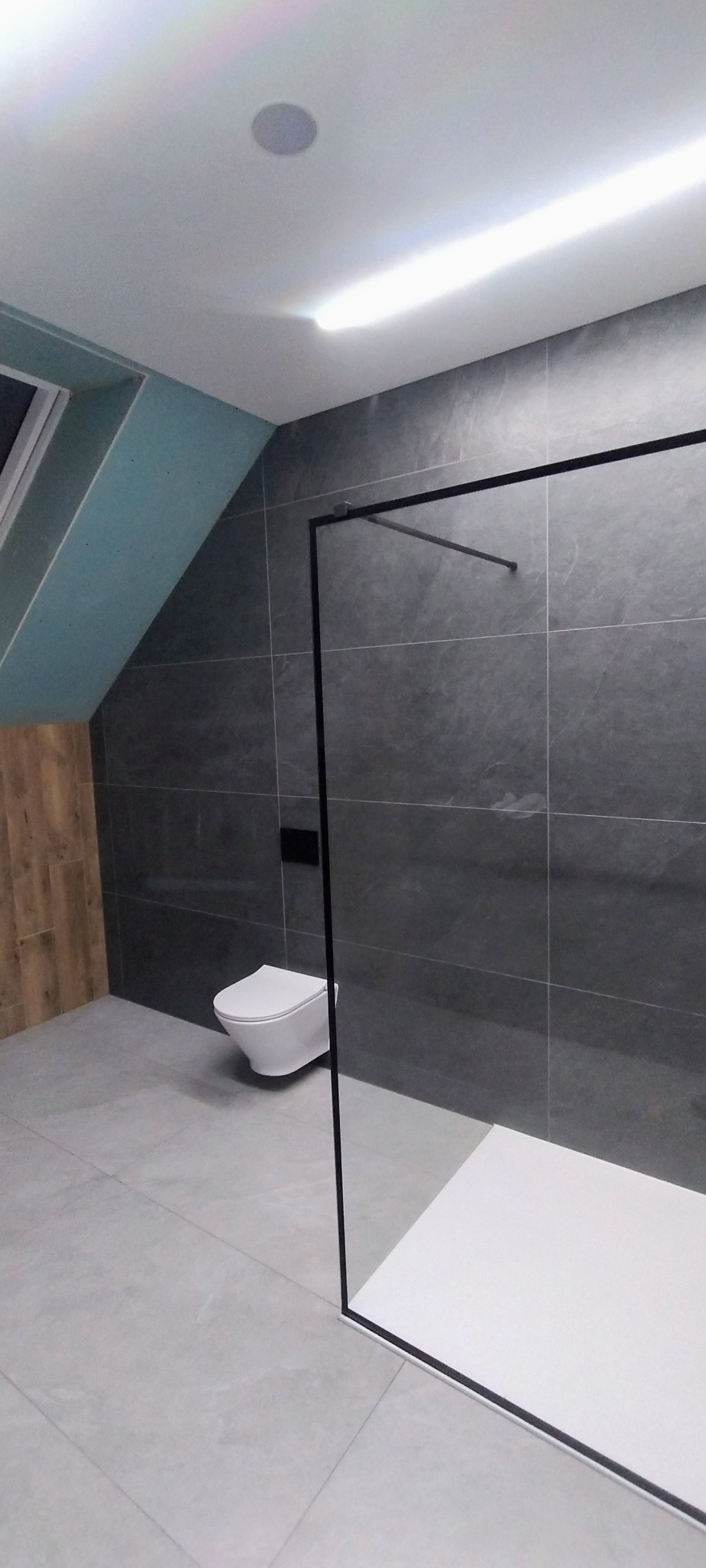 Nowoczesna łazienka na poddaszu z szarymi płytkami, kabiną prysznicową typu walk-in z czarną ramą i podwieszaną toaletą. Widoczne skosy i oświetlenie sufitowe.