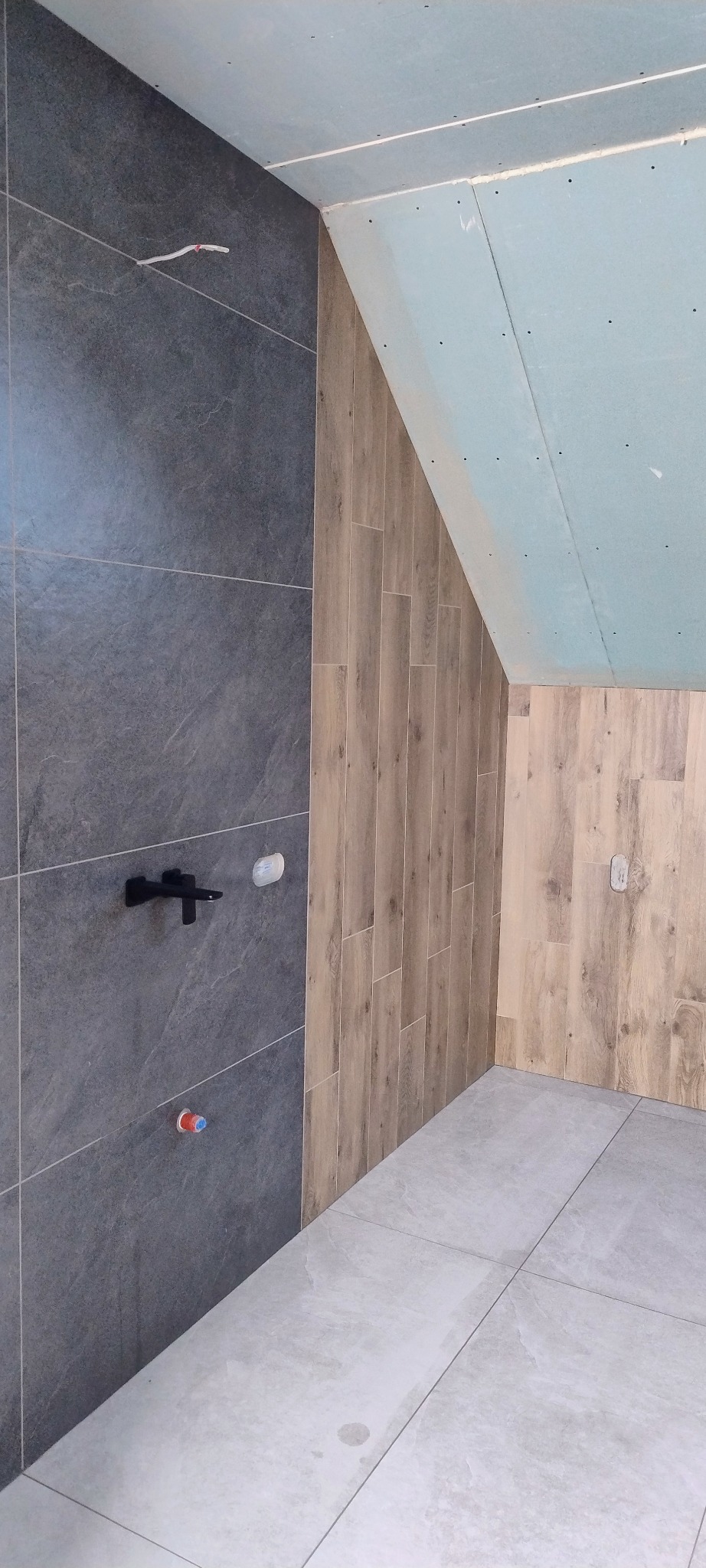 Wykończona łazienka w trakcie prac: szara płytka ścienna z czarną armaturą, drewnopodobne panele na skośnej ścianie poddasza, jasna podłoga z płytek.