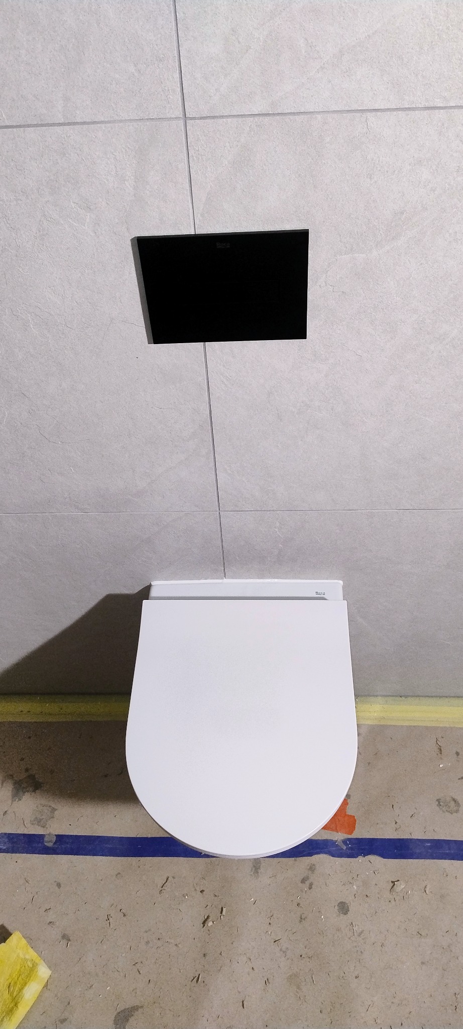 Biała, podwieszana miska WC marki Roca na tle szarych płytek i czarnego przycisku spłukującego. Widoczne zabezpieczenia podłogi. Wykończenie łazienki w minimalistycznym stylu.