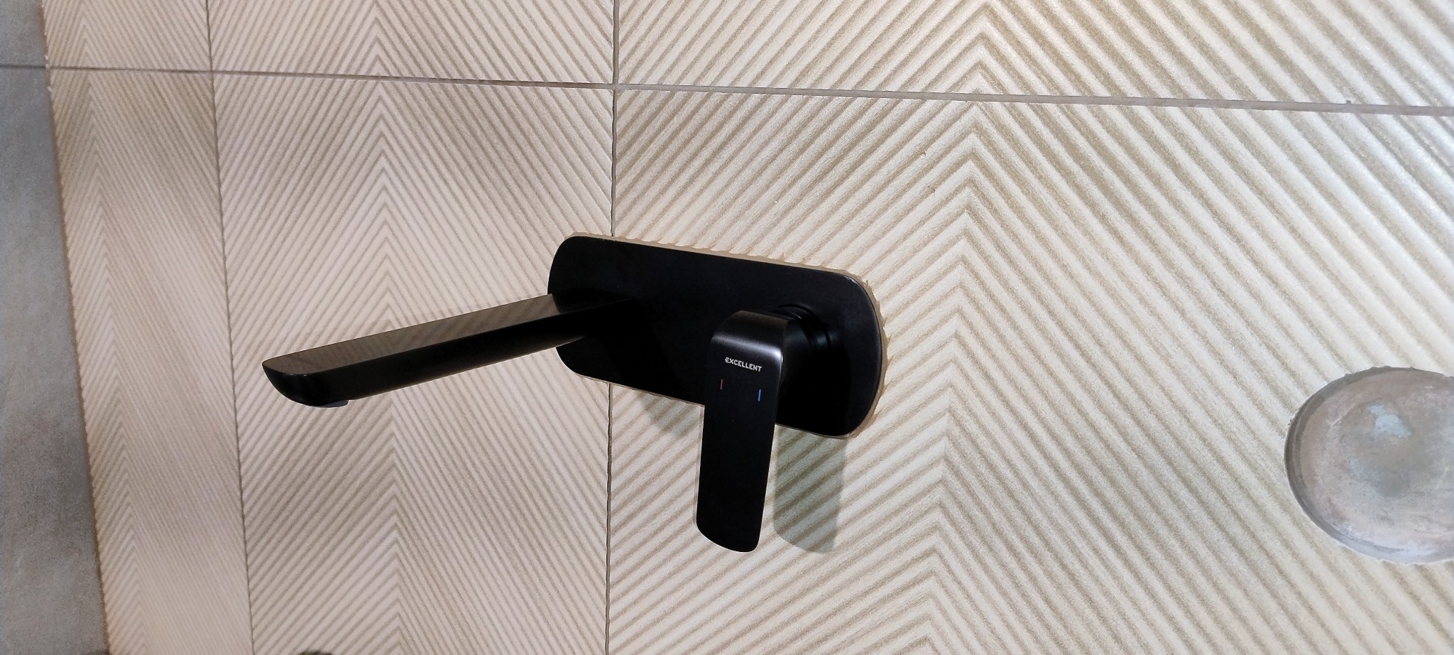 Czarna bateria umywalkowa marki Excellent na ścianie z wzorzystych, beżowych płytek w łazience. Widoczny otwór na dodatkowy element instalacji.