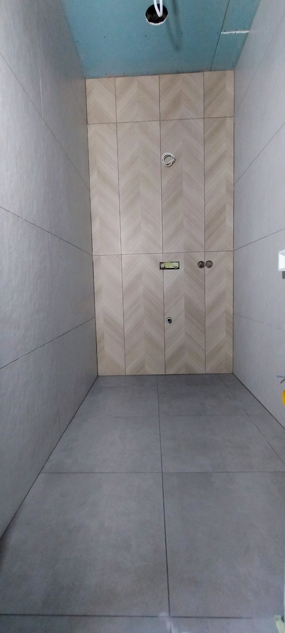 Wykończona łazienka z szarą podłogą i ścianami. Jedna ściana z płytek ułożonych w jodełkę. Widoczne otwory na gniazdka i instalacje. Sufit w kolorze niebieskim.