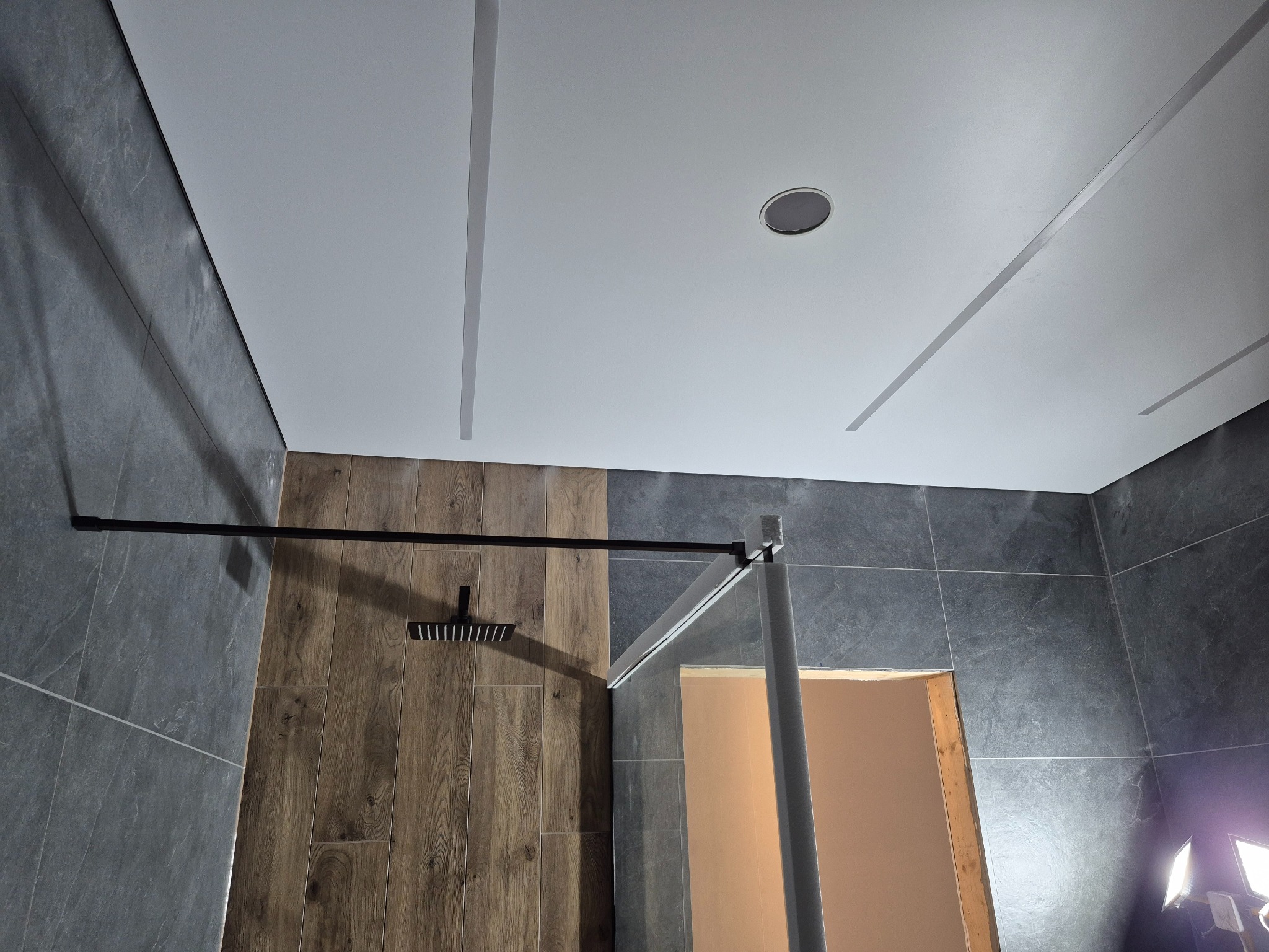 Nowoczesna kabina prysznicowa z czarną armaturą i szklaną ścianką, wykończona płytkami w odcieniach szarości i drewna. Oświetlenie sufitowe z liniowymi elementami.