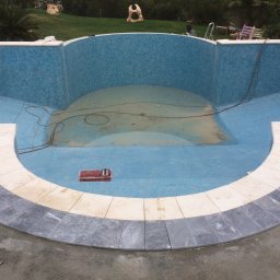 topKAwork - Niedokończony basen z mozaiką w odcieniach błękitu i szarości, widoczne narzędzia w czerwonym pojemniku, świeżo ułożone obrzeże z jasnego kamienia i szare płytki na zewnątrz basenu, trawa w tle.