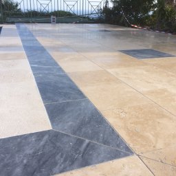 topKAwork - Wykończona posadzka z płytek w odcieniach beżu i szarości wokół basenu, z widocznym fragmentem niebieskiej wody i zieleni w tle.