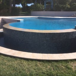 topKAwork - Niebieski basen z mozaikową obudową i kamiennym obrzeżem, otoczony zielonym trawnikiem i roślinnością.