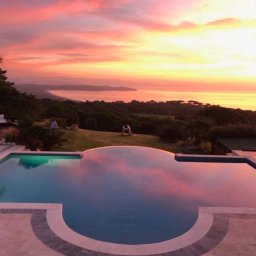 topKAwork - Infinity pool reflecting a vibrant sunset over a coastal landscape with lush greenery and distant mountains, featuring a palm tree and outdoor furniture.