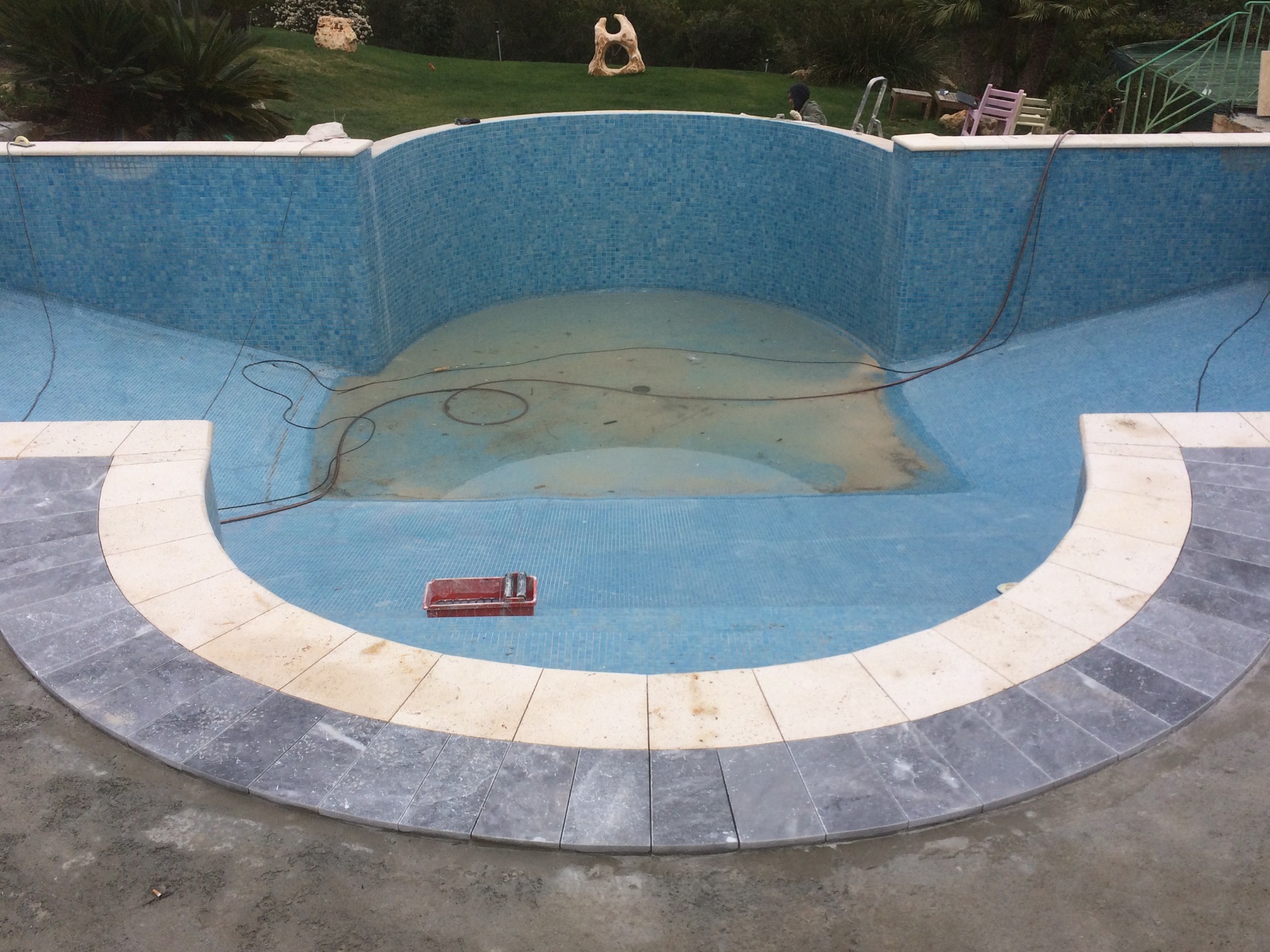 Niedokończony basen z mozaiką w odcieniach błękitu i szarości, widoczne narzędzia w czerwonym pojemniku, świeżo ułożone obrzeże z jasnego kamienia i szare płytki na zewnątrz basenu, trawa w tle.