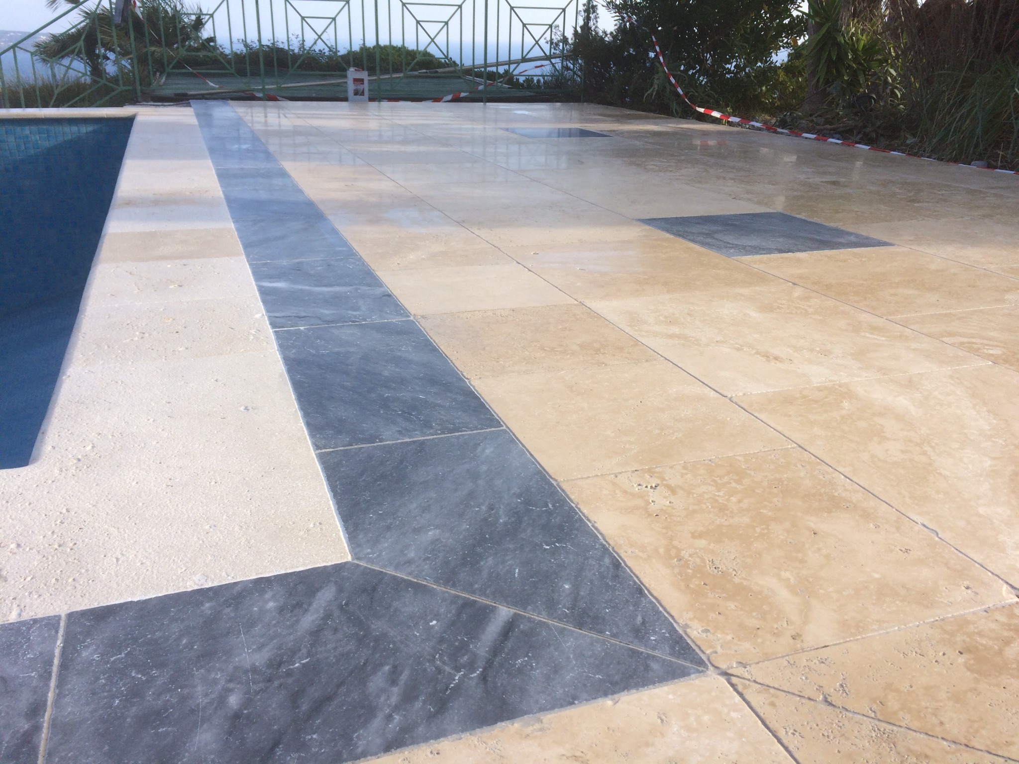 Wykończona posadzka z płytek w odcieniach beżu i szarości wokół basenu, z widocznym fragmentem niebieskiej wody i zieleni w tle.