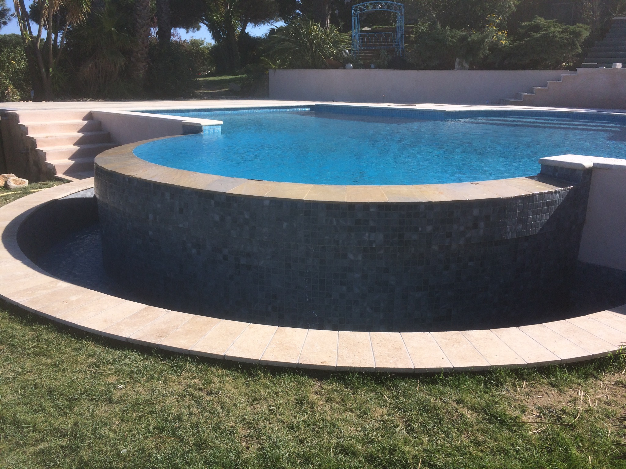 Niebieski basen z mozaikową obudową i kamiennym obrzeżem, otoczony zielonym trawnikiem i roślinnością.