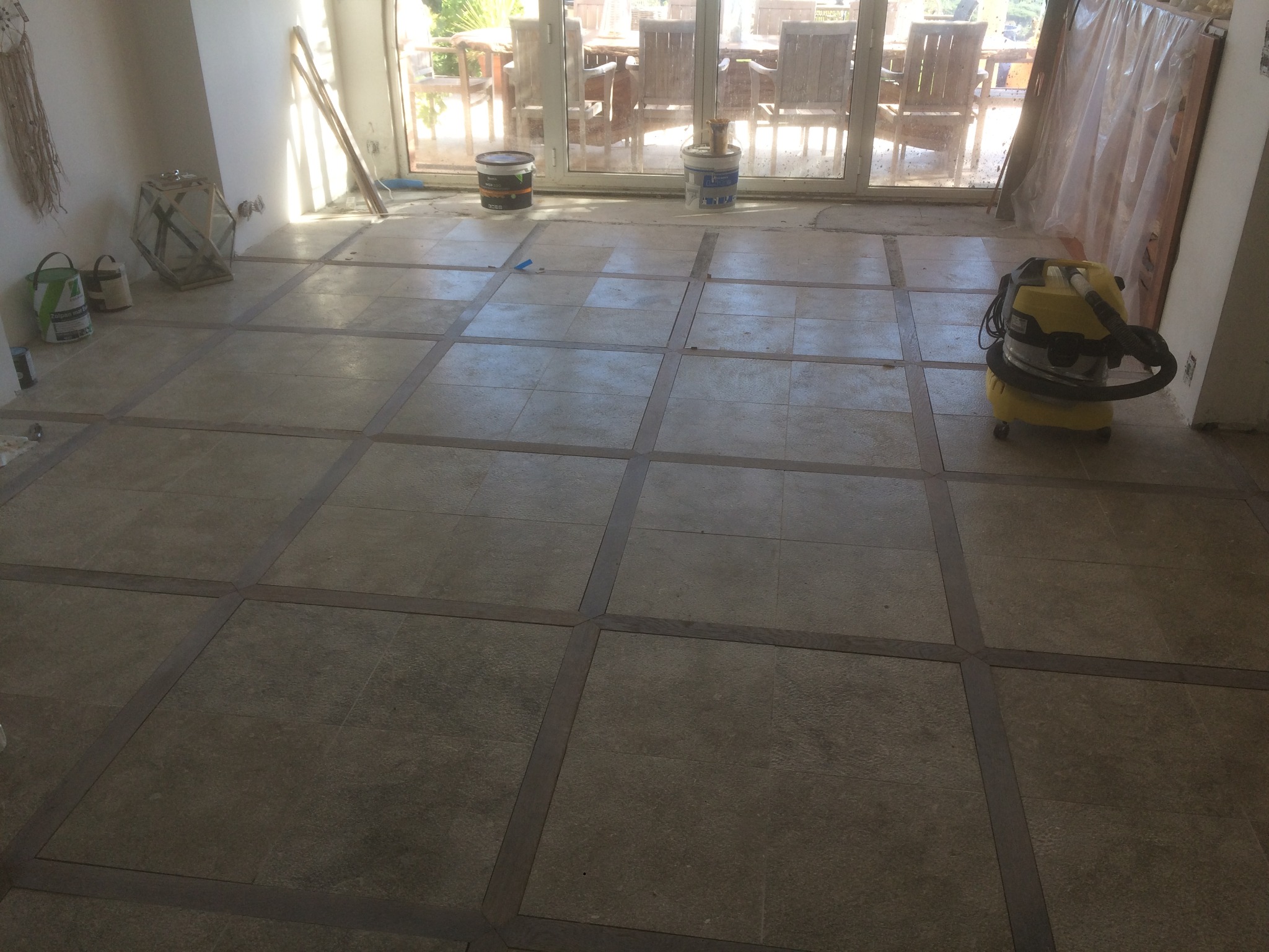 Układanie płytek podłogowych w kwadraty z ciemniejszymi obramowaniami, widoczne puszki z farbą, odkurzacz przemysłowy i narzędzia w tle.