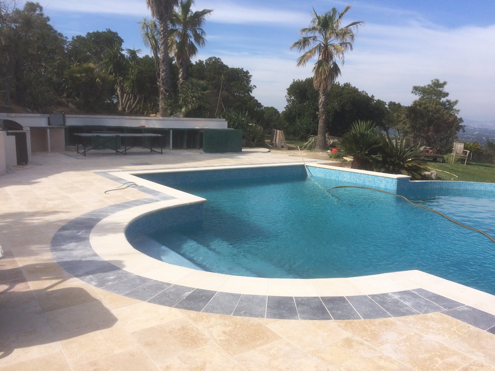 Niebieski basen z mozaiką i jasnobeżową kamienną obudową na zewnątrz, z palmami w tle i zabudowanym grillem z blatem roboczym.