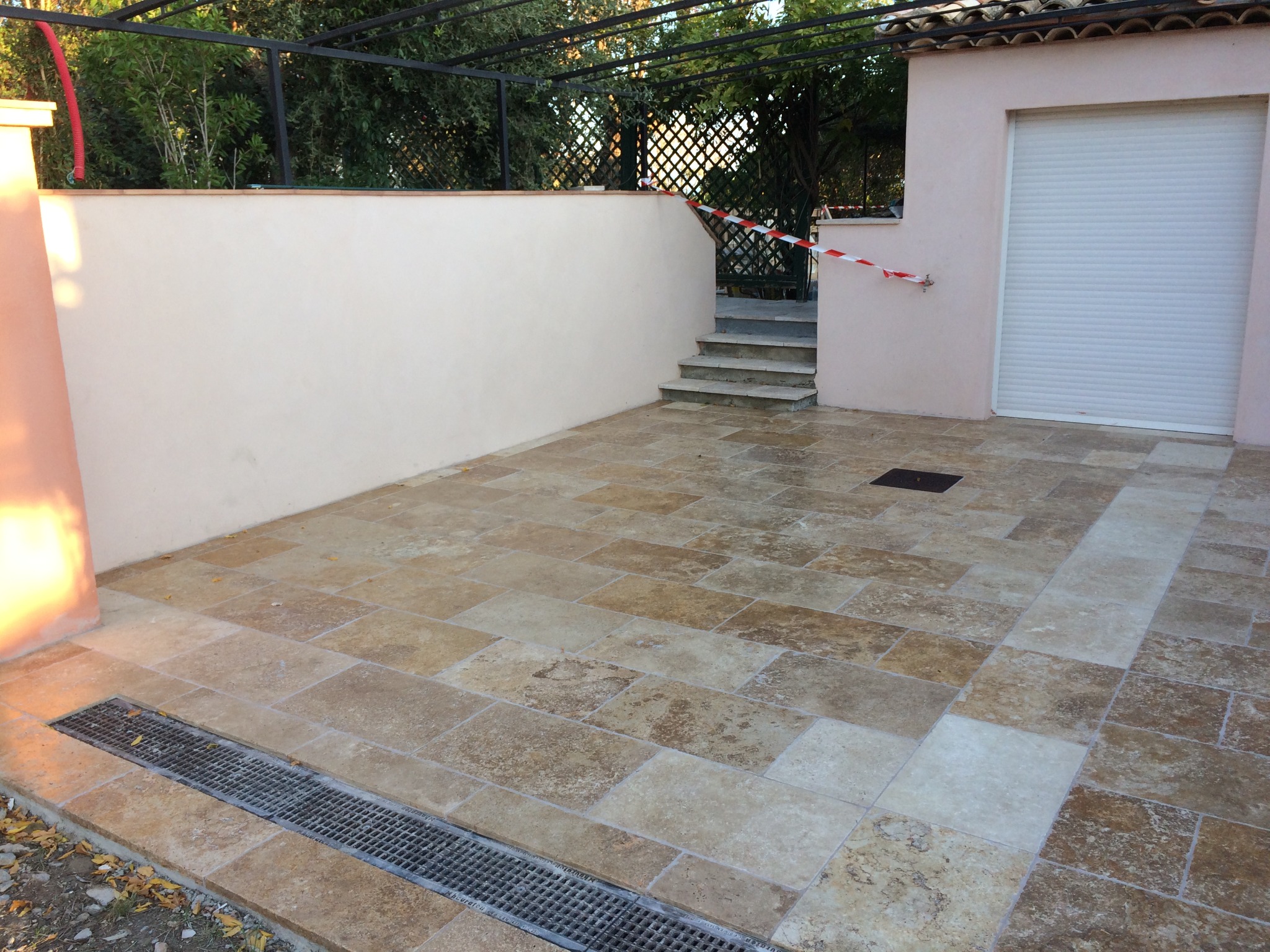 Wyłożony trawertynem taras zewnętrzny z odpływem liniowym, zabezpieczony taśmą ostrzegawczą przy schodach prowadzących do ogrodu.