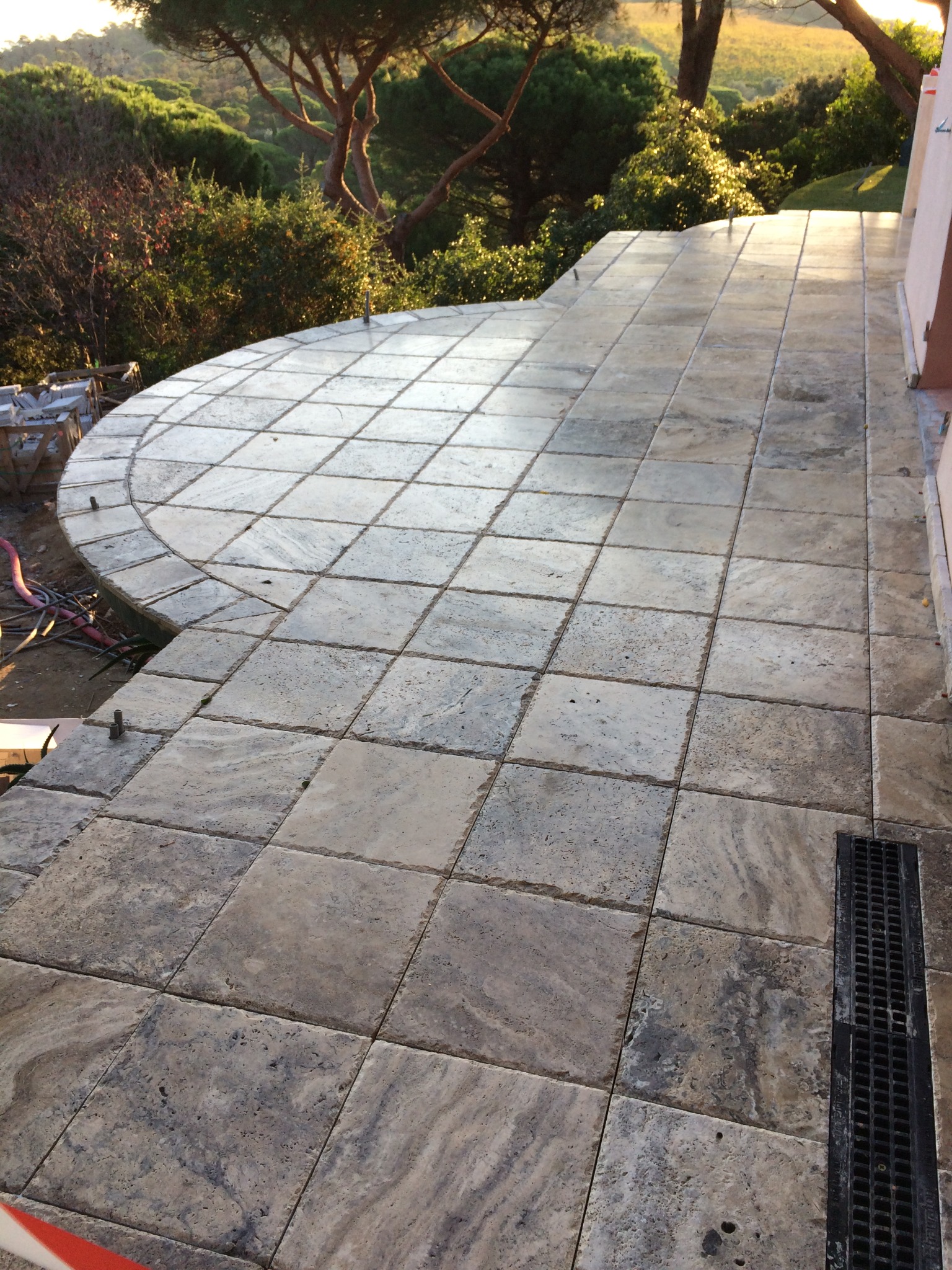 Ułożona kamienna posadzka tarasu z widocznym odpływem liniowym i fragmentem półokrągłego wykończenia, w tle zieleń i drzewa.