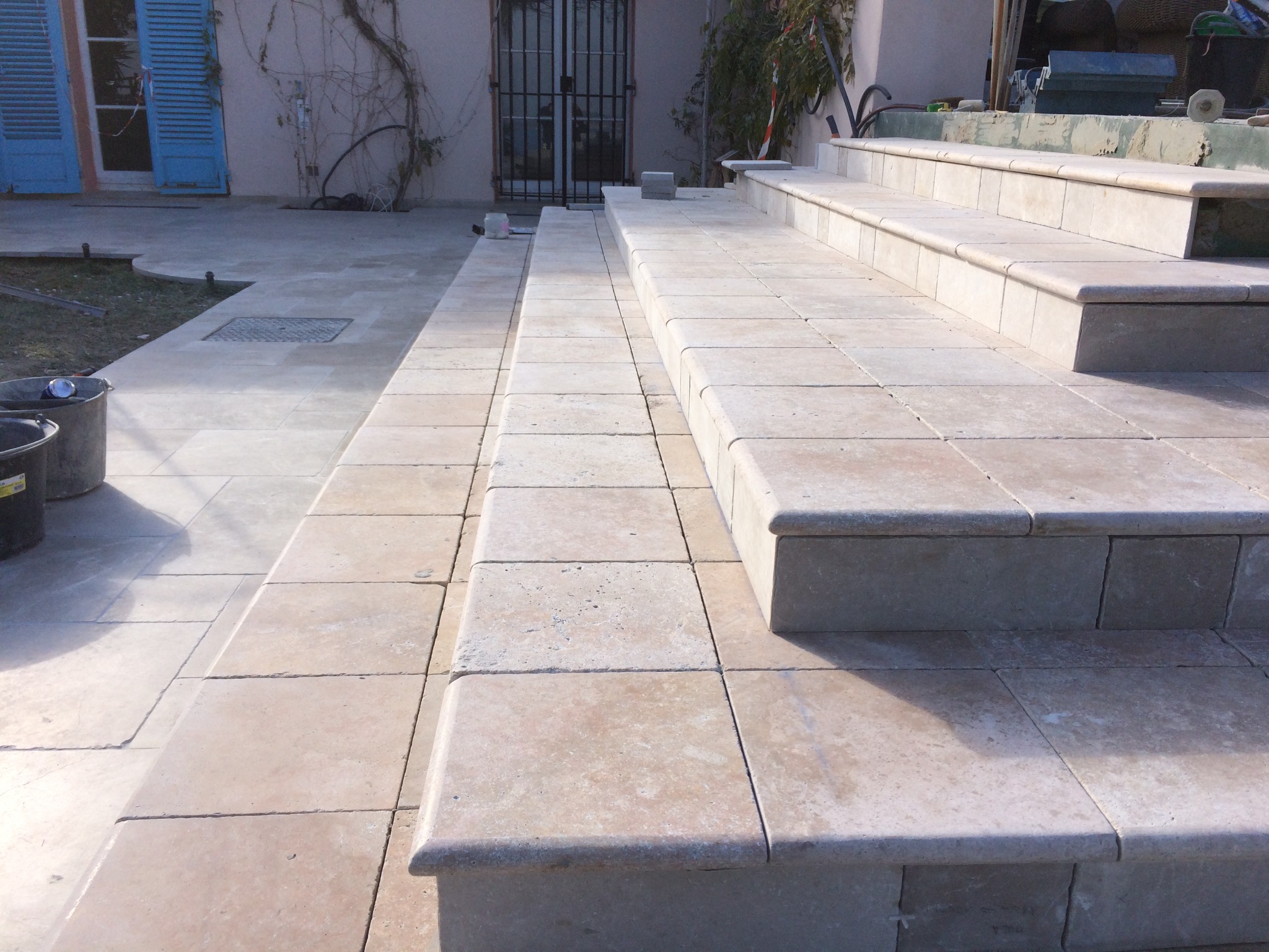 Wykonane z jasnego kamienia schody zewnętrzne z zaokrąglonymi krawędziami stopni, prowadzące do drzwi z metalową kratą, obok niebieskich okiennic.