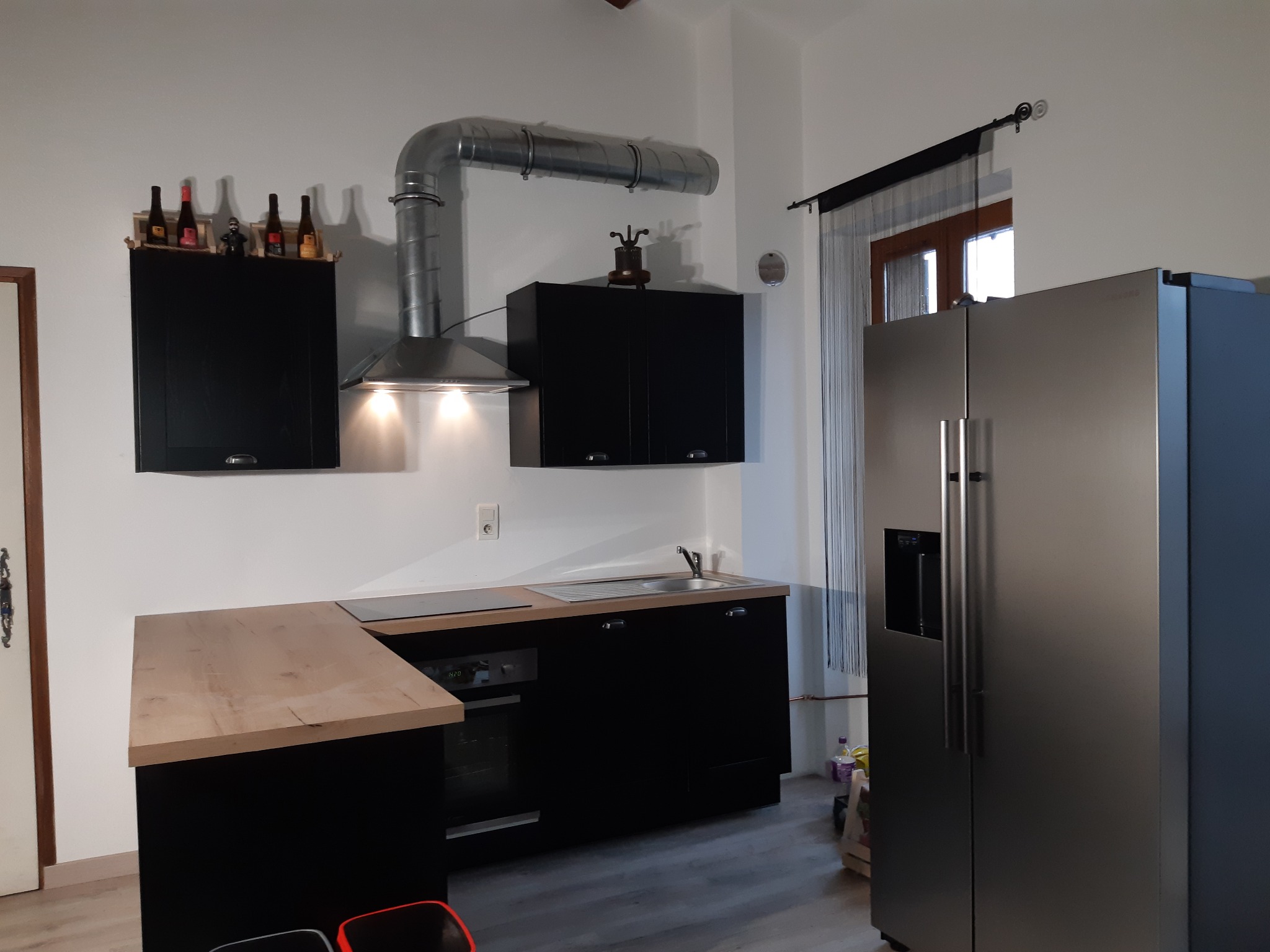 Nowoczesna kuchnia w czerni i drewnie: czarne szafki wiszące z dekoracyjnymi butelkami na górze, srebrny okap, drewniany blat i duża srebrna lodówka z dozownikiem wody.