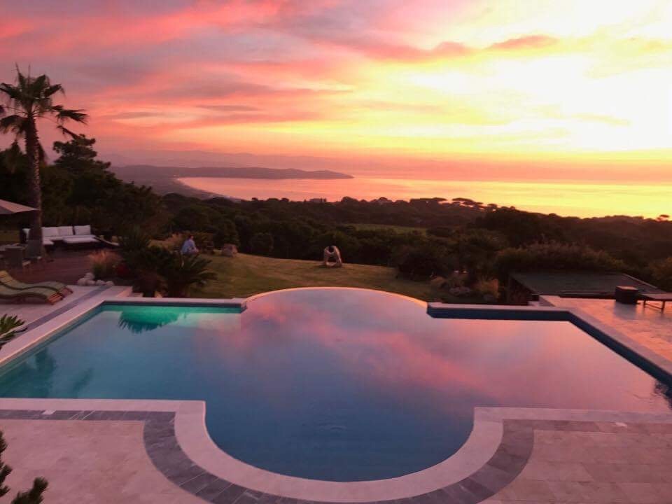 Infinity pool reflecting a vibrant sunset over a coastal landscape with lush greenery and distant mountains, featuring a palm tree and outdoor furniture.