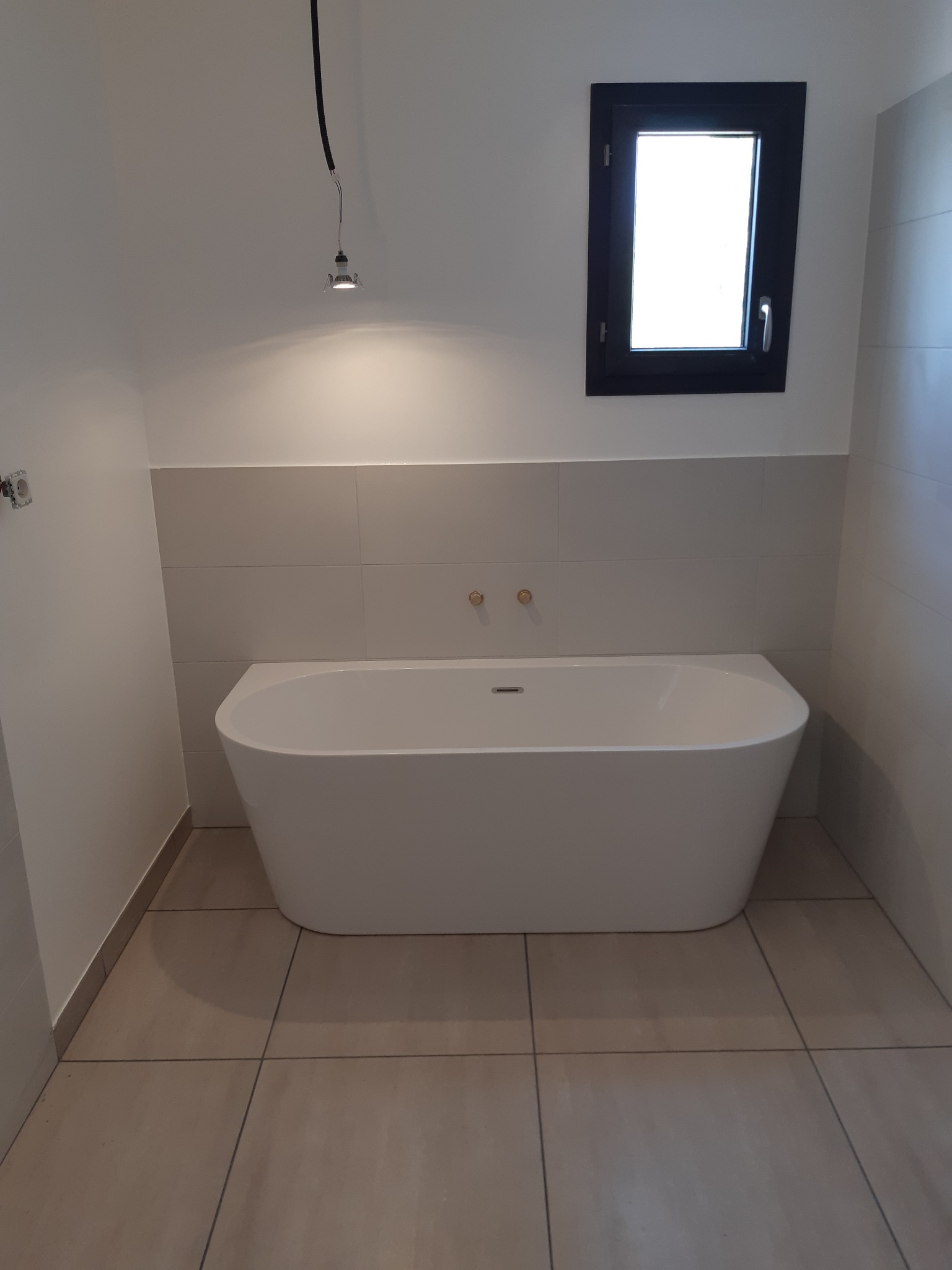 Nowoczesna wolnostojąca wanna w minimalistycznej łazience z beżowymi płytkami podłogowymi i białymi płytkami ściennymi, oświetlona wiszącą lampą punktową i naturalnym światłem z małego, czarnego okna.