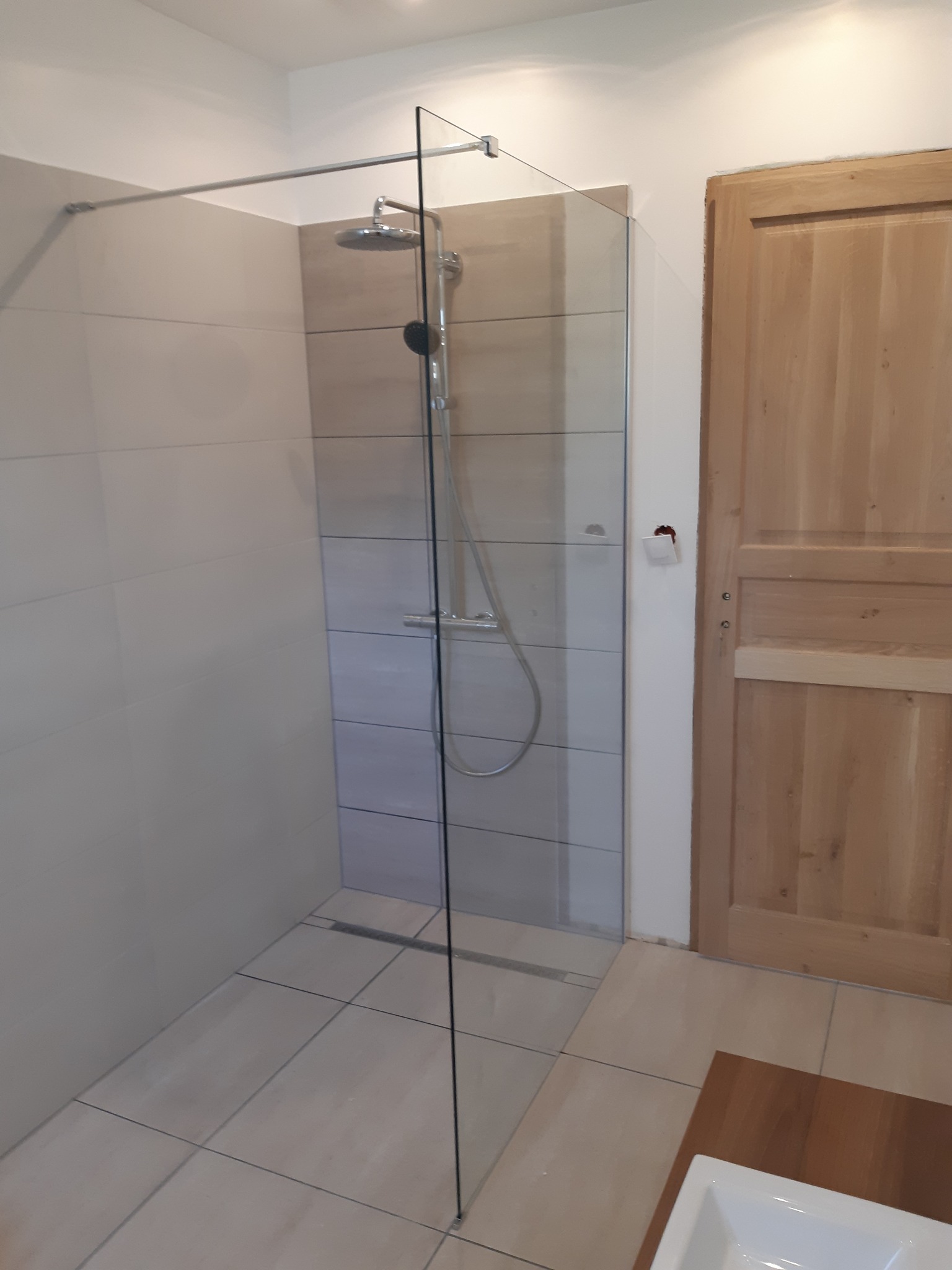 Nowoczesna kabina prysznicowa walk-in z transparentną szybą, zamontowana na jasnych płytkach w łazience z drewnianymi drzwiami i niedokończonym wykończeniem ściany wokół włącznika.