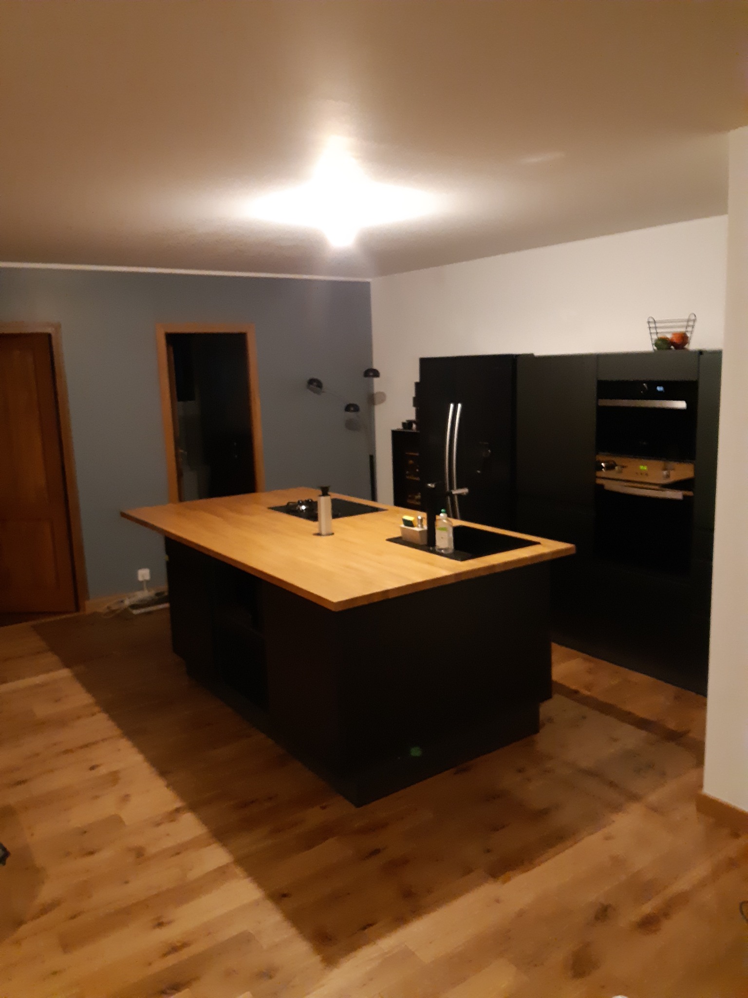 Nowoczesna kuchnia z wyspą o drewnianym blacie i czarnych szafkach, widoczne sprzęty AGD w zabudowie i drewniana podłoga.