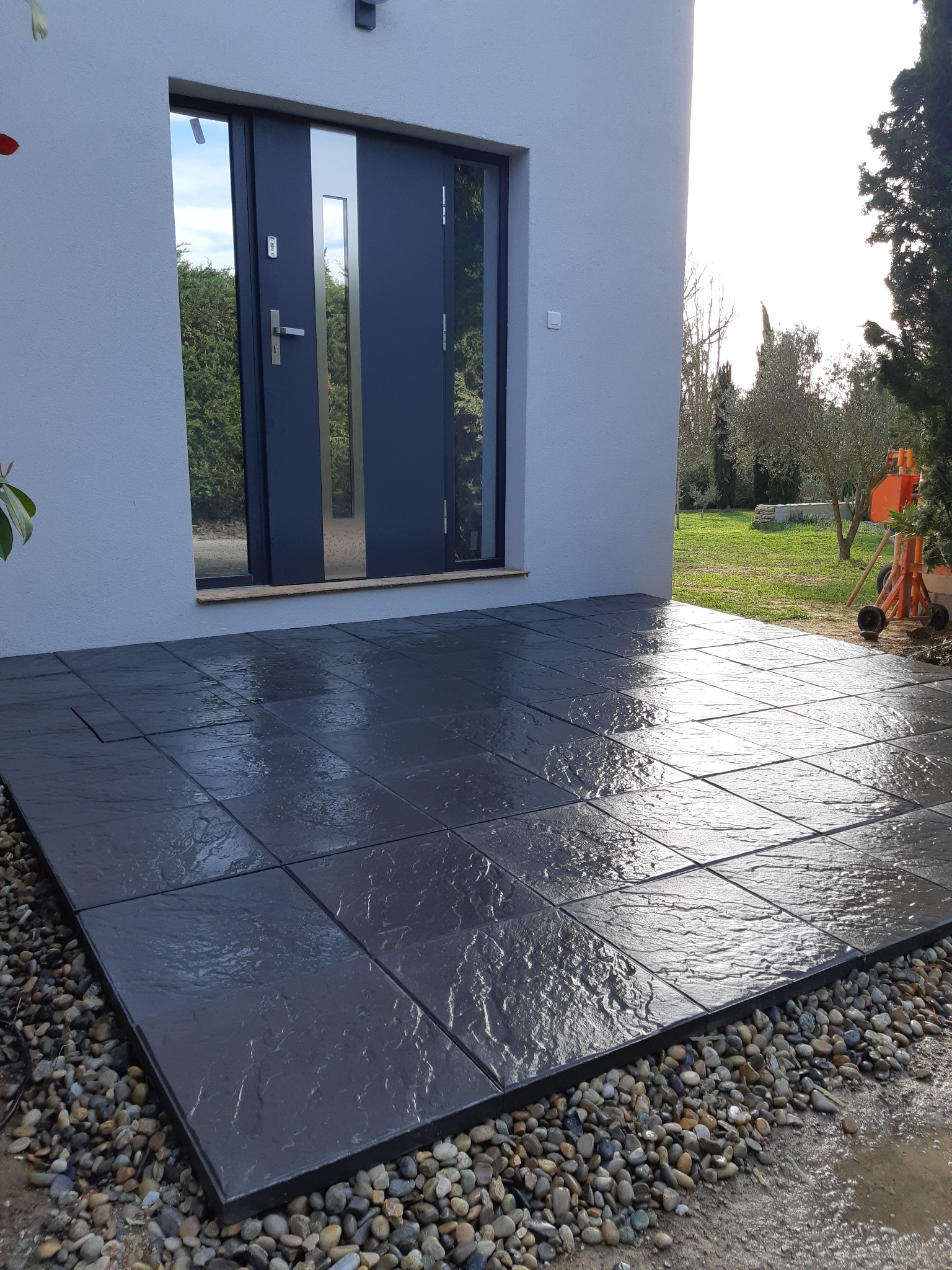 Nowoczesny taras z ciemnoszarych płyt kamiennych, wykończony drobnymi kamykami, przylegający do budynku z szarą elewacją i stalowymi drzwiami wejściowymi.