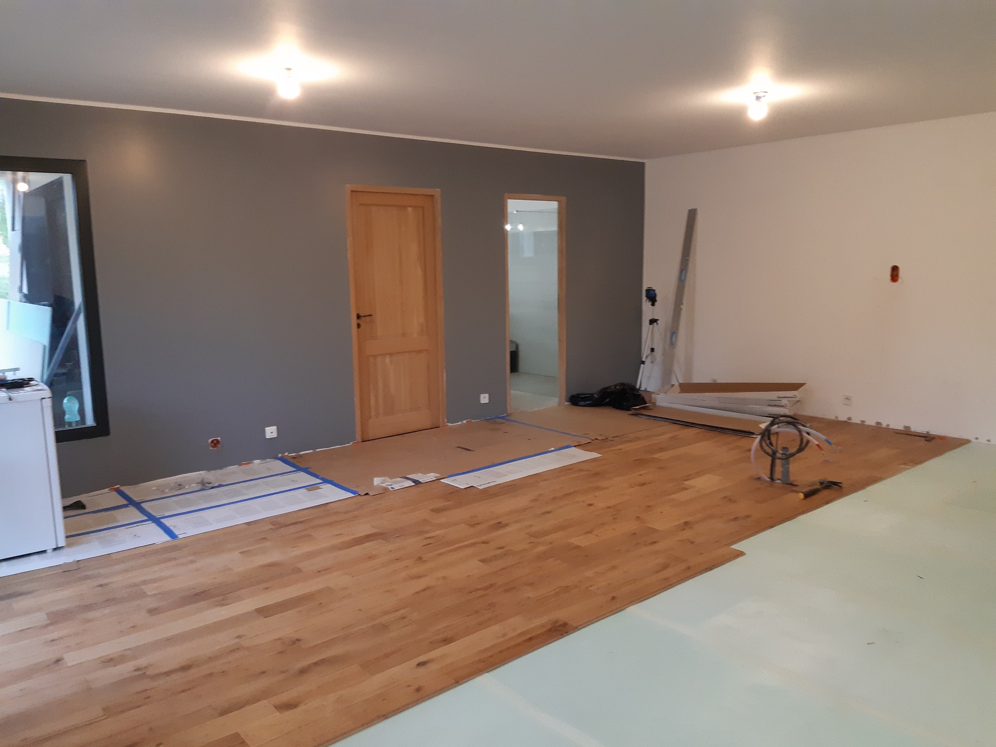 Układanie drewnianej podłogi w pokoju z szarą ścianą, widoczne narzędzia, niebieska taśma malarska zabezpieczająca podłoże i biała podkładka pod panele.