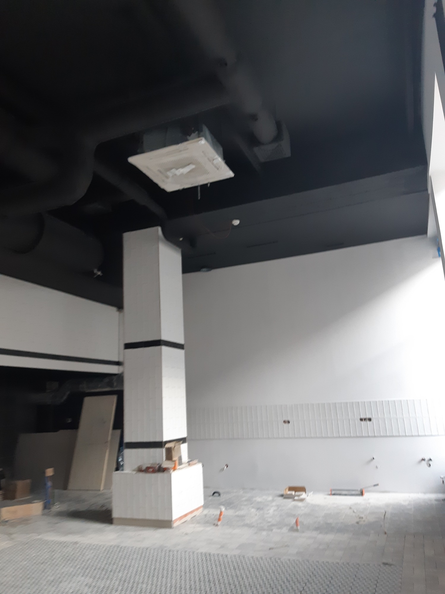 Wnętrze w trakcie wykańczania: białe płytki na ścianach i kolumnie, czarny sufit z widoczną instalacją, szara podłoga z wzorem, narzędzia i materiały budowlane porozrzucane po podłodze.