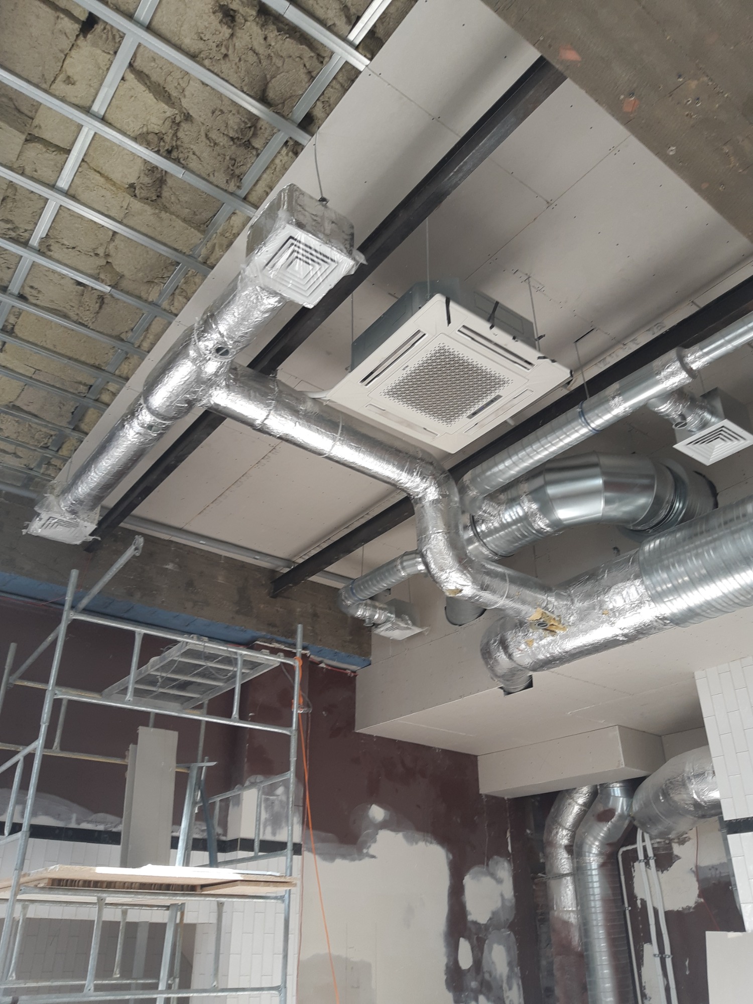 Montaż instalacji wentylacyjnej i klimatyzacji pod sufitem podwieszanym z płyt kartonowo-gipsowych w Lublinie, widoczne kanały wentylacyjne w izolacji, kratki nawiewne i rusztowanie.