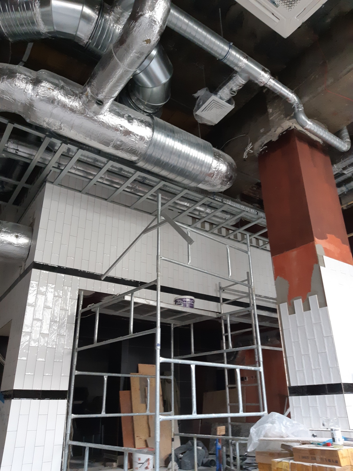 Montaż instalacji wentylacyjnej z metalowymi rurami pod sufitem, widoczne rusztowanie, częściowo wykończona ściana z białymi płytkami ceramicznymi i pomalowany na brązowo filar.