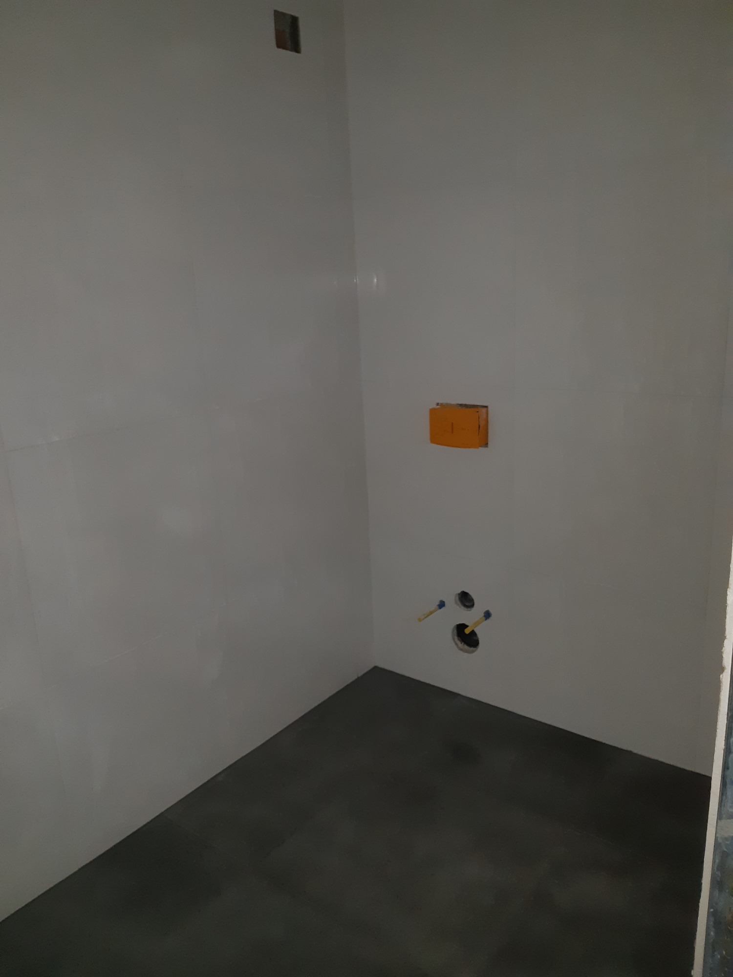 Przygotowanie do montażu armatury w wykończonej łazience z białymi płytkami ściennymi i ciemną podłogą, widoczne wystające rury i puszka instalacyjna.