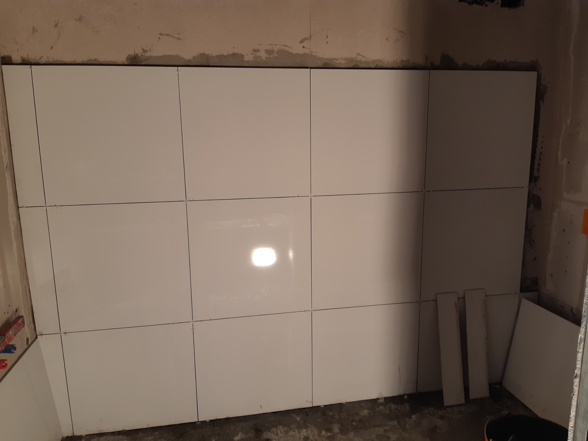 Ściana w trakcie układania białych, kwadratowych płytek z wąskimi fugami, widoczne krzyżyki dystansowe i fragment surowej ściany powyżej.