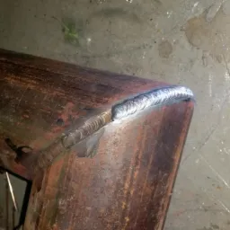 Spoina na zardzewiałym kolanku rury stalowej, widoczny spaw na łączeniu pod kątem prostym, w tle żółta tuba z logo ESAB.