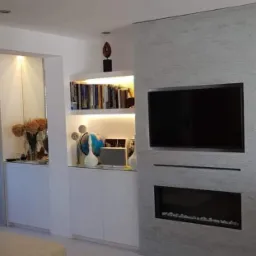 Nowoczesny salon z zabudową ściany telewizyjnej, w której znajduje się kominek elektryczny i półki z podświetleniem LED, widoczne dekoracje i książki.