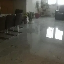 Błyszcząca, szara posadzka kamienna w salonie, odbijająca światło okienne i elementy wyposażenia, w tle stół z krzesłami, telewizor oraz rośliny doniczkowe.