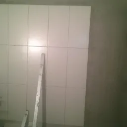 Ściana w trakcie wykańczania z ułożonymi białymi płytkami, widoczna drabina i poziomice, surowe ściany dookoła.