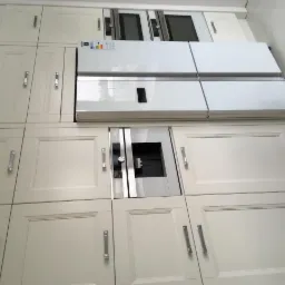 Jasna zabudowa kuchenna z frezowanymi frontami, zintegrowana z lodówką side-by-side i sprzętem AGD, widoczna z perspektywy pod kątem.