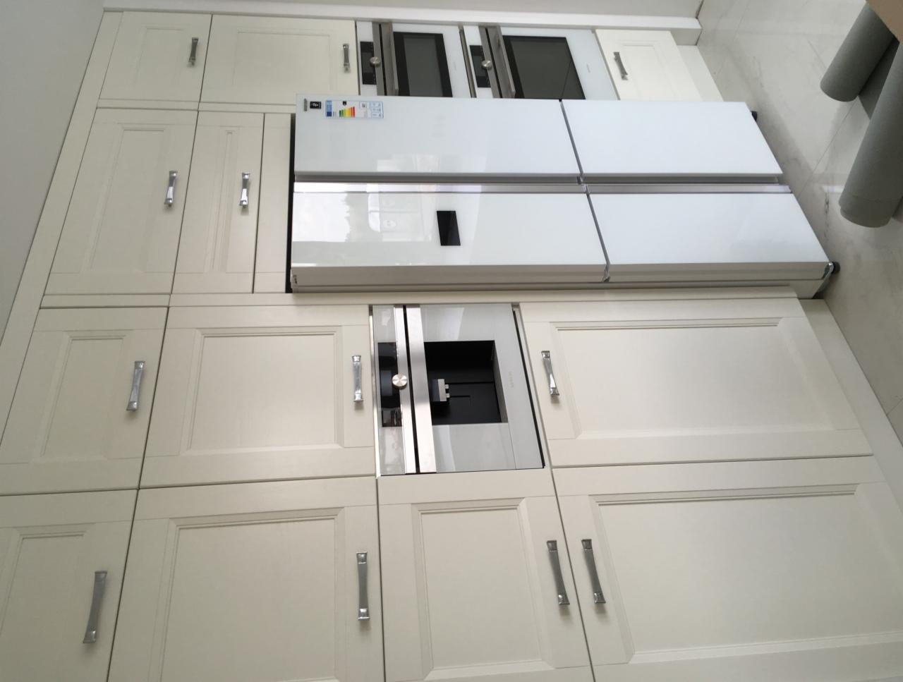 Jasna zabudowa kuchenna z frezowanymi frontami, zintegrowana z lodówką side-by-side i sprzętem AGD, widoczna z perspektywy pod kątem.