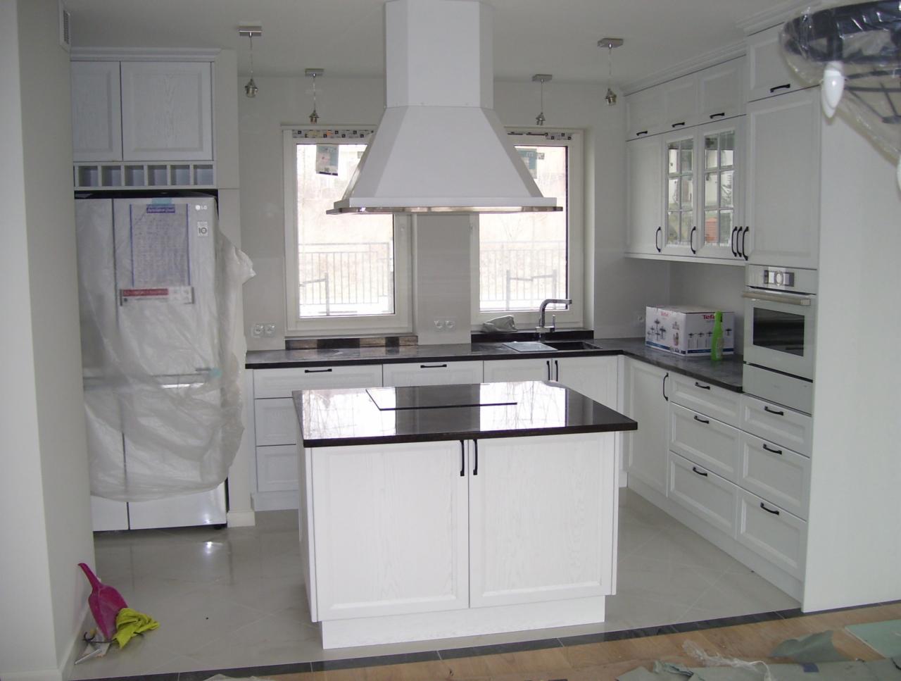 Jasna kuchnia z białą zabudową i czarnym blatem, wyspa kuchenna na pierwszym planie, lodówka zabezpieczona folią ochronną, widoczne drobne niedociągnięcia wykończeniowe.