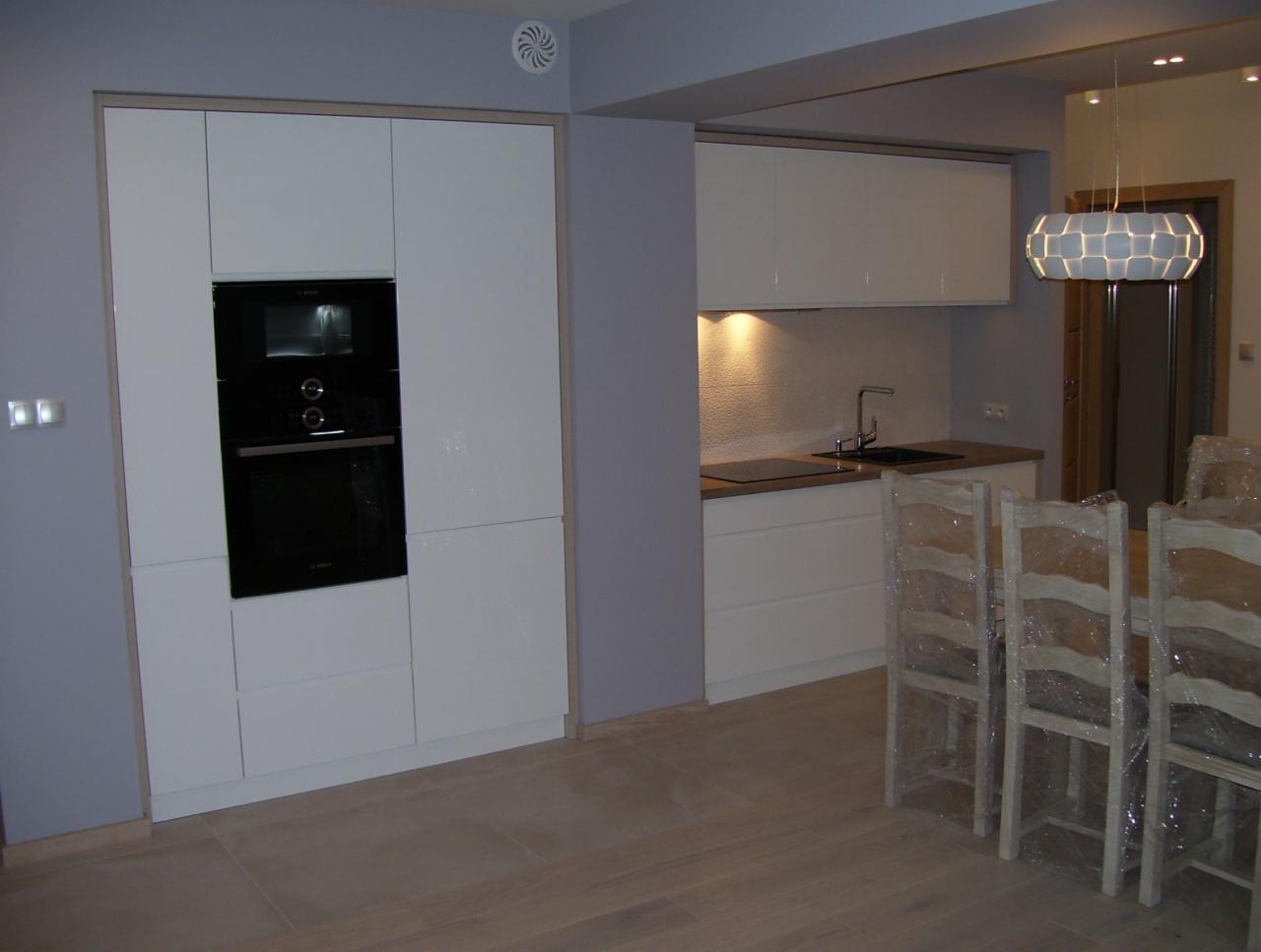 Nowoczesna kuchnia w odcieniach bieli i szarości, z zabudową kuchenną z piekarnikiem i płytą grzewczą, oświetlona wiszącą lampą, krzesła owinięte folią ochronną.