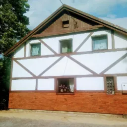 Budynek z muru pruskiego po renowacji, z ceglaną podstawą, widoczne trzy okna na górze i dwa na dole, otoczenie zieleni i częściowo zachmurzone niebo.