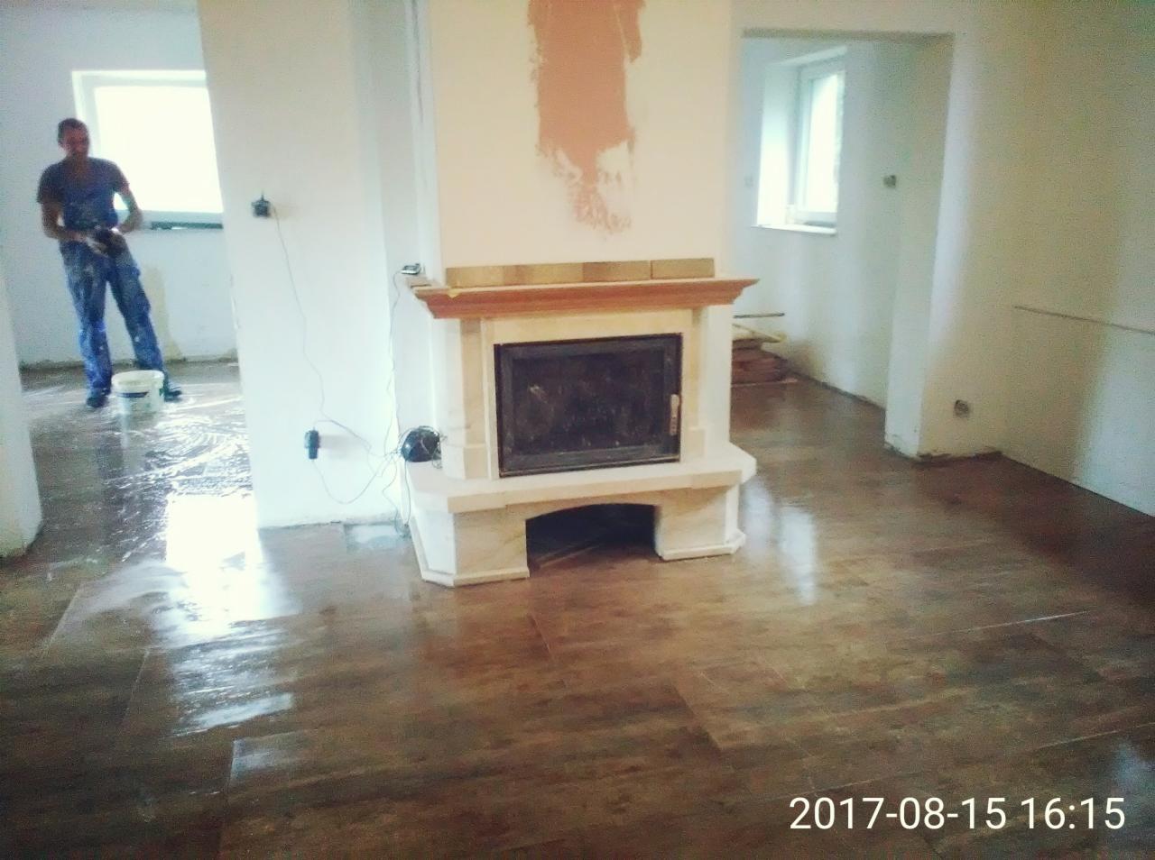 Wnętrze w trakcie remontu: świeżo położona, błyszcząca podłoga imitująca drewno, biały kominek z drewnianymi elementami, ściany częściowo zagruntowane, w tle majster w niebieskim kombinezonie...