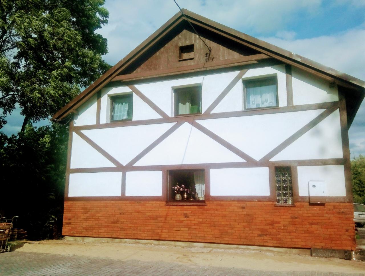 Budynek z muru pruskiego po renowacji, z ceglaną podstawą, widoczne trzy okna na górze i dwa na dole, otoczenie zieleni i częściowo zachmurzone niebo.