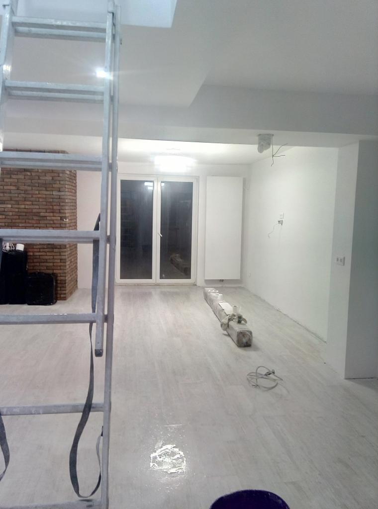 Wnętrze pokoju po remoncie z widoczną drabiną malarską, zakrytym podłożem, świeżo pomalowanymi ścianami i ceglanym kominkiem w tle.